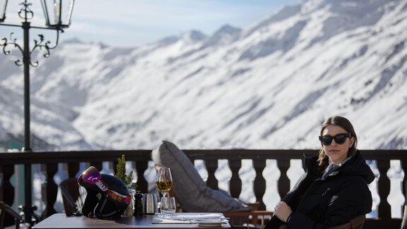 Eine Frau sitzt entspannt an einem Tisch in einer Berghütte. Im Hintergrund sind schneebedeckte Berge und ein klarer Himmel zu sehen. - TOP Hotel Hochgurgl - Hochgurgl