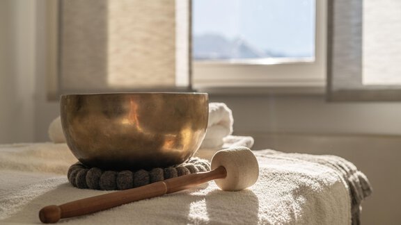 Eine Klangschale aus Metall steht auf einem hellen Tisch mit einem Hammer daneben. Im Hintergrund sind Fenster mit sanftem Licht sichtbar. - TOP Hotel Hochgurgl - Hochgurgl