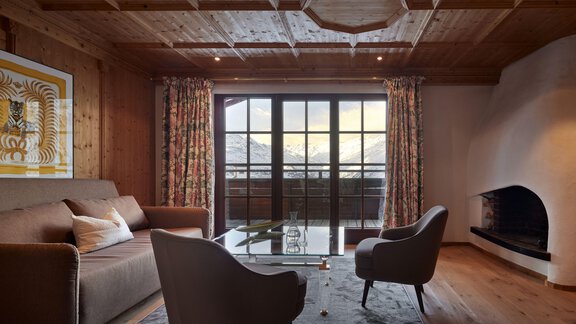 Ein gemütlicher Wohnraum mit Holzwänden und einem großen Fenster, das einen Blick auf die Berge bietet. Die Einrichtung besteht aus einem Sofa, zwei Sesseln und einem eleganten Glastisch. - TOP Hotel Hochgurgl - Hochgurgl
