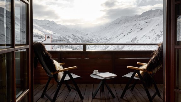 Ein gemütlicher Balkon mit zwei Stühlen und einem Tisch, der auf eine schneebedeckte Berglandschaft blickt. Die Wolken verdecken teilweise die Sonne, was eine ruhige Atmosphäre schafft. - TOP Hotel Hochgurgl - Hochgurgl