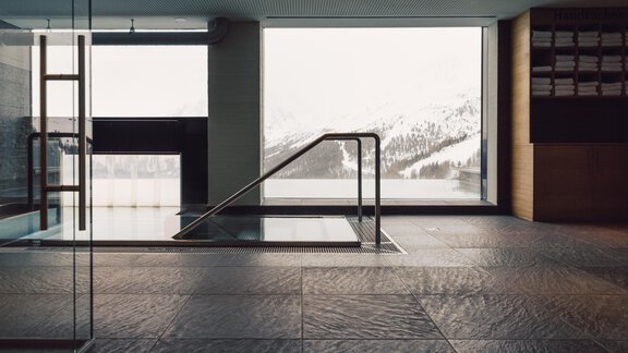 An elegant wellness area with a pool and floor-to-ceiling windows. In the background, snow-covered mountains and a tranquil lake can be seen. - 4 * Superior Hotel Schöne Aussicht - Hochsölden