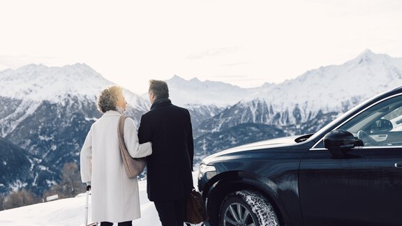 A couple stands on a snow-covered mountain, surrounded by majestic peaks. Next to them, a black car is parked as they enjoy the view. - 4 * Superior Hotel Schöne Aussicht - Hochsölden