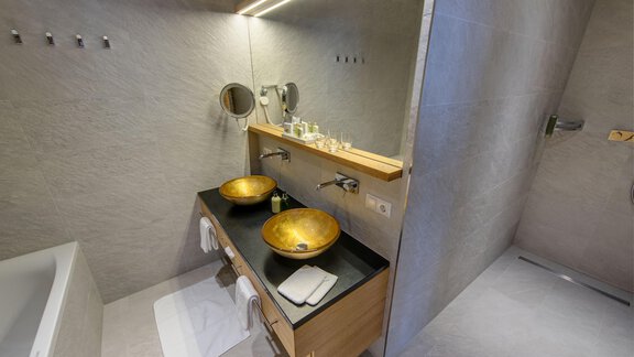 A modern bathroom with two golden sinks and a large mirror. The walls are in neutral colors, and there is a shower as well as a bathtub. - 4 * Superior Hotel Schöne Aussicht - Hochsölden