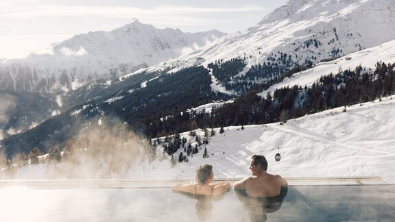 A couple relaxes in a heated outdoor pool with a view of snowy mountains. The scene radiates tranquility and well-being. - 4 * Superior Hotel Schöne Aussicht - Hochsölden