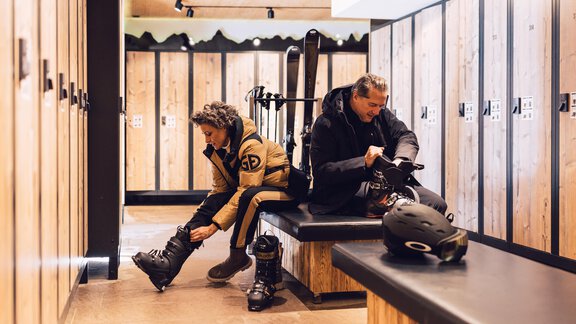 A ski area with wooden lockers, where two people are putting on their ski gear. One person is sitting on a bench putting on their ski boots, while the other is focused on the ground. - 4 * Superior Hotel Schöne Aussicht - Hochsölden