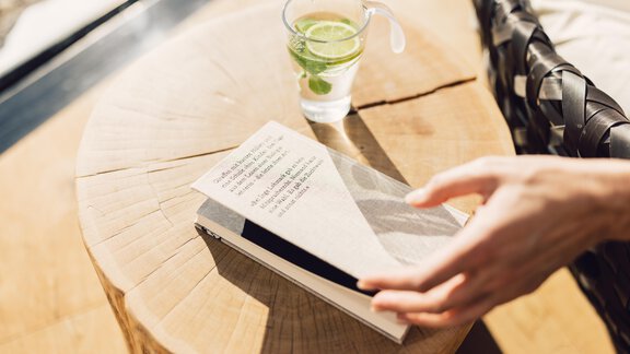 A hand with a book on a wooden table. Next to the book is a glass with a drink and a lime slice. - 4 * Superior Hotel Schöne Aussicht - Hochsölden