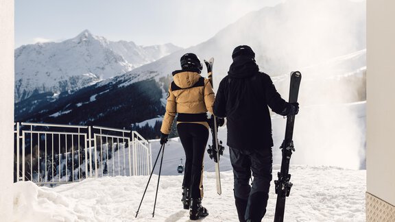 A couple is walking on a snow-covered area while carrying their skis. Majestic mountains and blue sky can be seen in the background. - 4 * Superior Hotel Schöne Aussicht - Hochsölden