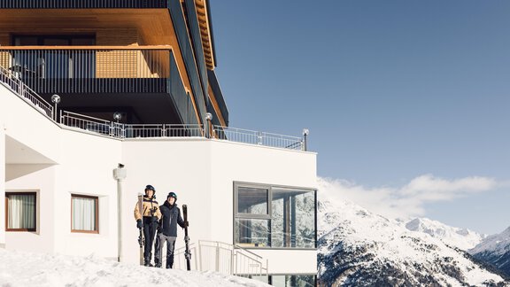A modern lodge in the snow with a view of the mountains. Two skiers are standing in front of the building and enjoying the alpine landscape. - 4 * Superior Hotel Schöne Aussicht - Hochsölden
