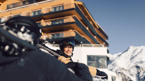 A laughing female skier wearing a helmet carries her skis near a modern mountain chalet. In the background, snow-covered mountains and a clear sky can be seen. - 4 * Superior Hotel Schöne Aussicht - Hochsölden