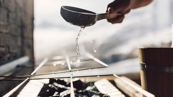 A hand holds a ladle over a grill area, from which water is splashing. In the background, a blurred landscape can be seen. - 4 * Superior Hotel Schöne Aussicht - Hochsölden