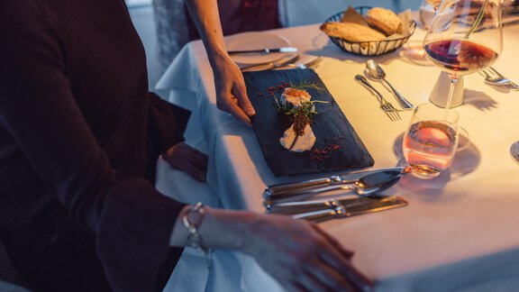 An elegant restaurant setting with a beautifully arranged plate on a table. A person is reaching for the plate, while glasses and cutlery are placed around them. - 4 * Superior Hotel Schöne Aussicht - Hochsölden