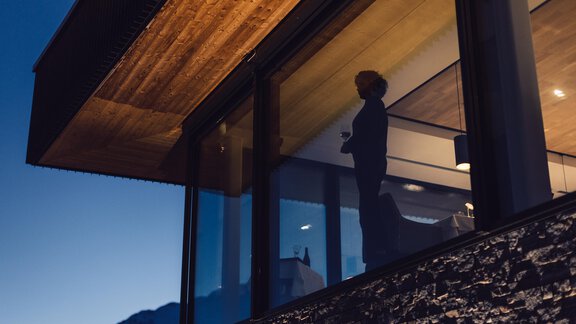 A modern apartment with large windows in the evening light. A person stands by the window and looks outside. - 4 * Superior Hotel Schöne Aussicht - Hochsölden