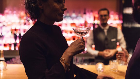 An elegantly dressed woman holds a cocktail glass in a stylish bar. In the background, the bartender is visible, surrounded by colorful drinks. - 4 * Superior Hotel Schöne Aussicht - Hochsölden