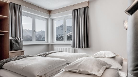 A cozy bedroom with two beds and large windows. Snow-capped mountains can be seen in the background. - Enzian Apartments - Hochsölden