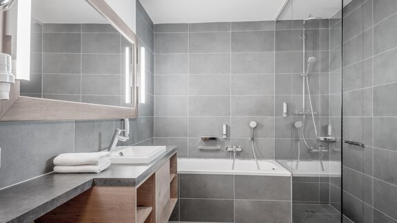 A modern bathroom with gray tiles and a bathtub. The wooden sink cabinet and the mirror lights give the room an elegant touch. - Enzian Apartments - Hochsölden