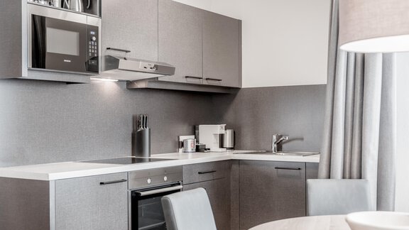 A modern kitchen with gray cabinets and stainless steel appliances. There is a microwave, a stove, and an elegant dining table. - Enzian Apartments - Hochsölden