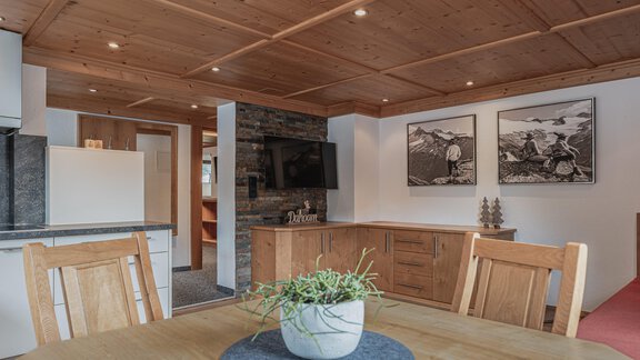 A stylish living room with wooden furnishings and a modern kitchen unit. The walls are decorated with pictures and a television, and a table with a plant stands in the foreground. - Appartements Alpenrose - Huben
