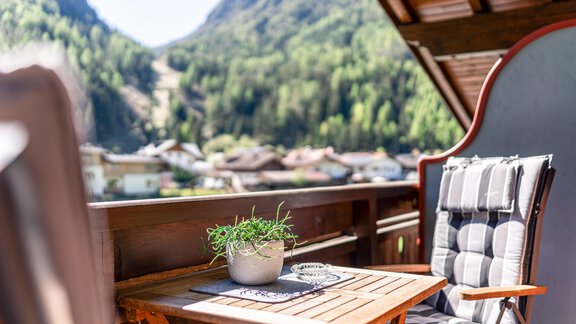 A cozy balcony with a table, a flowering plant, and a chair. In the background, green mountains and houses can be seen. - Appartements Alpenrose - Huben
