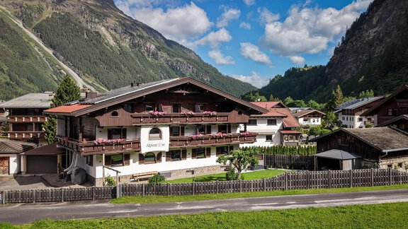 A traditional chalet in the Alps with blooming balconies. The surrounding landscape features impressive mountains and a green meadow. - Appartements Alpenrose - Huben