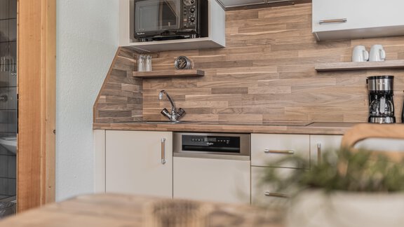 A modern kitchen with wood paneling and silver appliances. The room features a microwave oven, a coffee machine, and shelf space. - Appartements Alpenrose - Huben