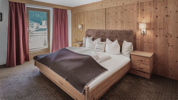 A cozy bedroom with a large bed and wooden walls. In front of the window, red curtains hang, creating a pleasant atmosphere. - Appartements Alpenrose - Huben