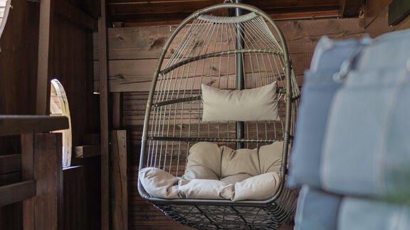 A beautiful hanging chair made of rattan with cushions can be seen in the room. The wooden ceiling and the walls give the room a cozy atmosphere. - Appartements Alpenrose - Huben