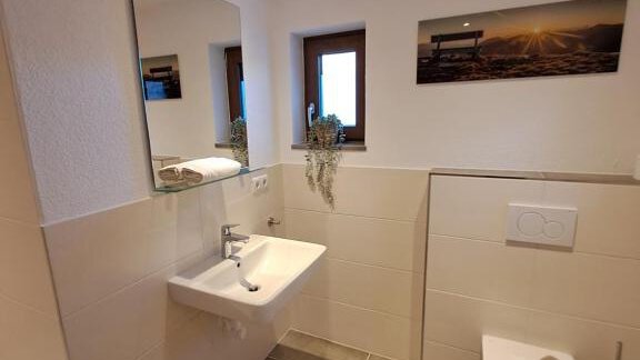 A modern bathroom with bright tiles and a large wall mirror. There is a sink and a toilet, as well as a window for natural light. - Berghaus Praxmarer - Huben