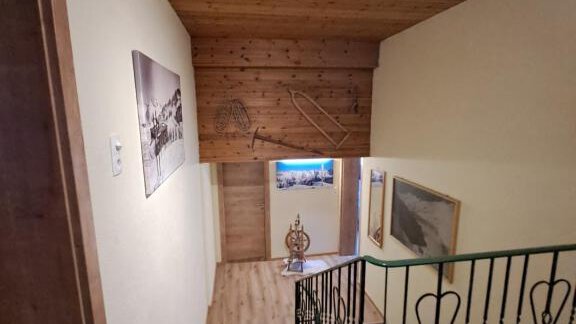 A staircase with a wooden ceiling and pleasant lighting. In the background, pictures are visible on the wall and a wooden door. - Berghaus Praxmarer - Huben