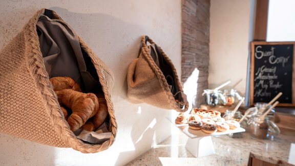 Two hanging baskets with fresh croissants on a wall. On a table, there are more sweet pastries and glasses. - Genusshotel Alpenblick - Huben