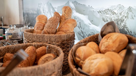 A buffet with various types of bread in baskets. In the background, snow-covered mountains can be seen. - Genusshotel Alpenblick - Huben