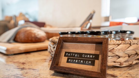 On a kitchen table lies a wooden frame with the inscription "Date Curry Spread". In the background, some bread and a few jars are visible. - Genusshotel Alpenblick - Huben
