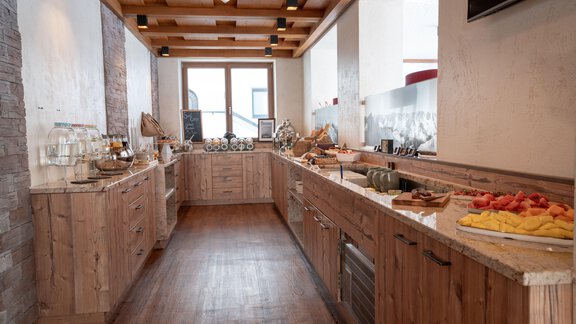 A modern kitchen with wooden cabinets and a long counter. On the counter are fresh fruits and various dishes arranged. - Genusshotel Alpenblick - Huben