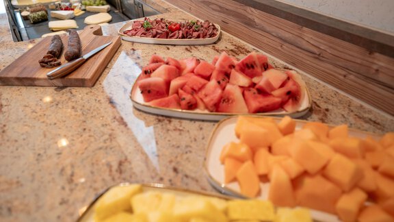A selection of fresh fruit, including watermelons, melons, and pineapples, is on a marbled table. In the background, cold cuts and other treats are visible. - Genusshotel Alpenblick - Huben