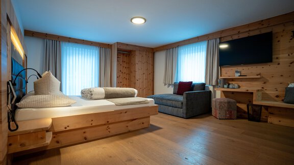 A cozy room with wooden furniture and large windows. There is a bed, a sofa, and a television. - Genusshotel Alpenblick - Huben