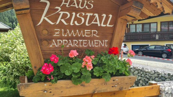 A wooden sign with the inscription "House Crystal" and colorful flowers. In the background, buildings and trees can be seen. - Haus Kristall - Huben