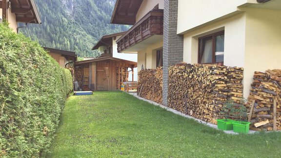 Eine grüne Wiese neben zwei Häusern, die mit Holzstapeln und einer Hecke umgeben sind. Im Hintergrund sind Berge zu sehen. - Haus Max - Huben