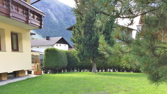 Ein grüner Garten mit Bäumen und einer Bergkulisse im Hintergrund. Im Vordergrund sieht man ein Wohnhaus und gepflegte Sträucher. - Haus Max - Huben