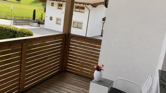 Ein gemütlicher Balkon aus Holz mit zwei weißen Stühlen und einem kleinen Tisch. Im Hintergrund sind grüne Hügel und einige traditionelle Häuser zu sehen. - ONO Apartments - Huben