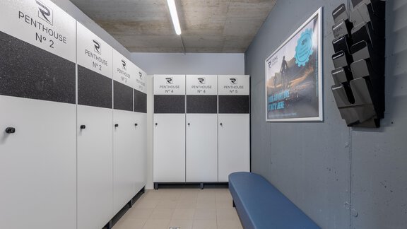 Ein moderner Umkleideraum mit weißen Schränken und einer blauen Bank. An der Wand hängen Informationen und eine Anzeige. - Alpenappartement Central Valley Penthouse - Längenfeld