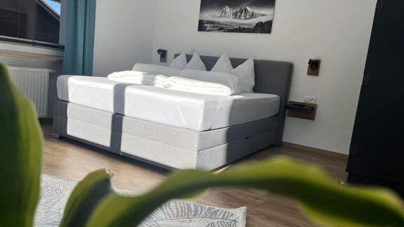 A modern bedroom with a large bed and elegant bed linens. In the background, there are windows and a piece of art on the wall. - Apart Aurelia - Längenfeld