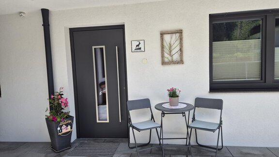 Ein Eingang mit einer modernen Tür und einem kleinen Tisch mit zwei Stühlen. Neben dem Tisch steht ein Blumentopf mit bunten Blumen. - Apart Fiegl Längenfeld - Längenfeld