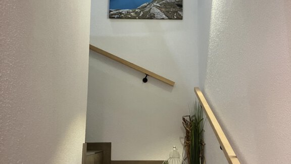 A bright staircase with a wooden railing leads up. On the wall hangs a picture of a blue lake and the surrounding area. - Apart Katja - Längenfeld