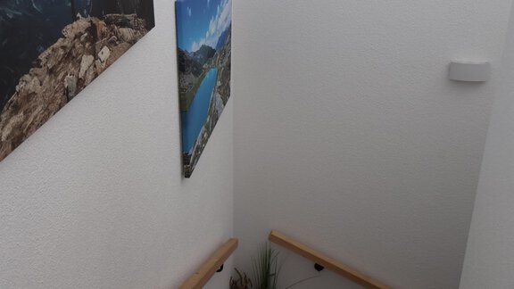 A staircase with a wooden railing and white walls leads downwards. Pictures of mountains and landscapes hang on the walls. - Apart Katja - Längenfeld