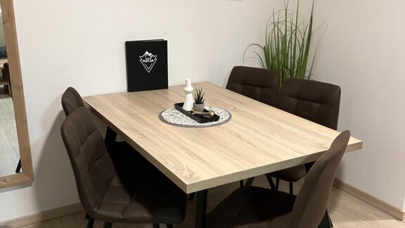 A modern dining area with a wooden table and six brown chairs. A television is mounted on the wall, and in the background, there is a plant. - Apart Katja - Längenfeld