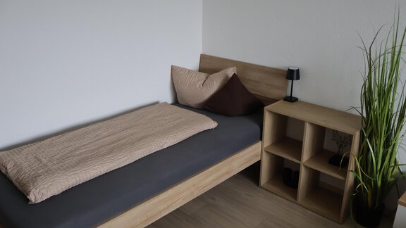 A simple room with a bed, a nightstand, and a green plant. The walls are light and the floor is made of wood. - Apart Katja - Längenfeld