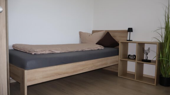 A simple bedroom with a wooden bed and a small bedside table.  
Next to the bed is a plant and a clock hangs on the wall. - Apart Katja - Längenfeld