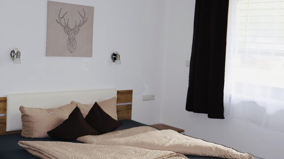A modern bedroom with a wooden bed and cozy bedding. The walls are painted light, and a large window provides plenty of daylight. - Apart Katja - Längenfeld