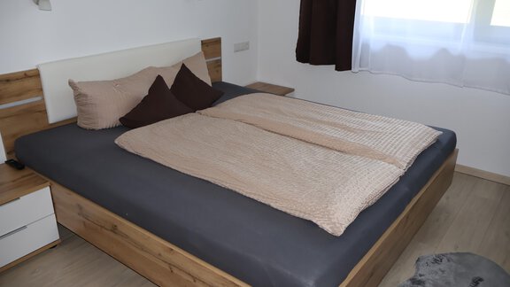 A modern bedroom with a wooden bed frame and a comfortable duvet. The walls are light, and there is a large window with curtains. - Apart Katja - Längenfeld