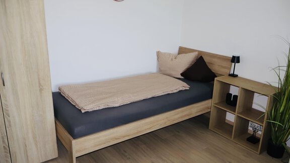 A simple bedroom with a bed, a small nightstand, and a plant. The walls are light and the floor is made of wood. - Apart Katja - Längenfeld