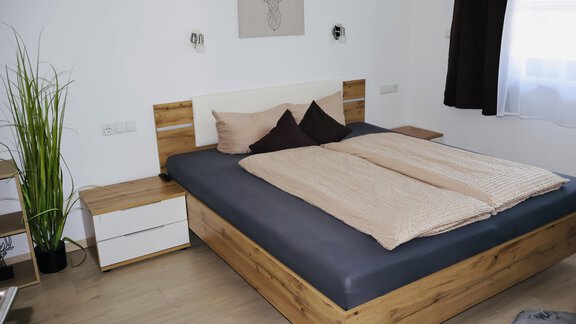 A cozy bedroom with a large wooden bed and beige bedcovers. On the wall hangs a picture of a deer antler and there is a green plant in the corner. - Apart Katja - Längenfeld